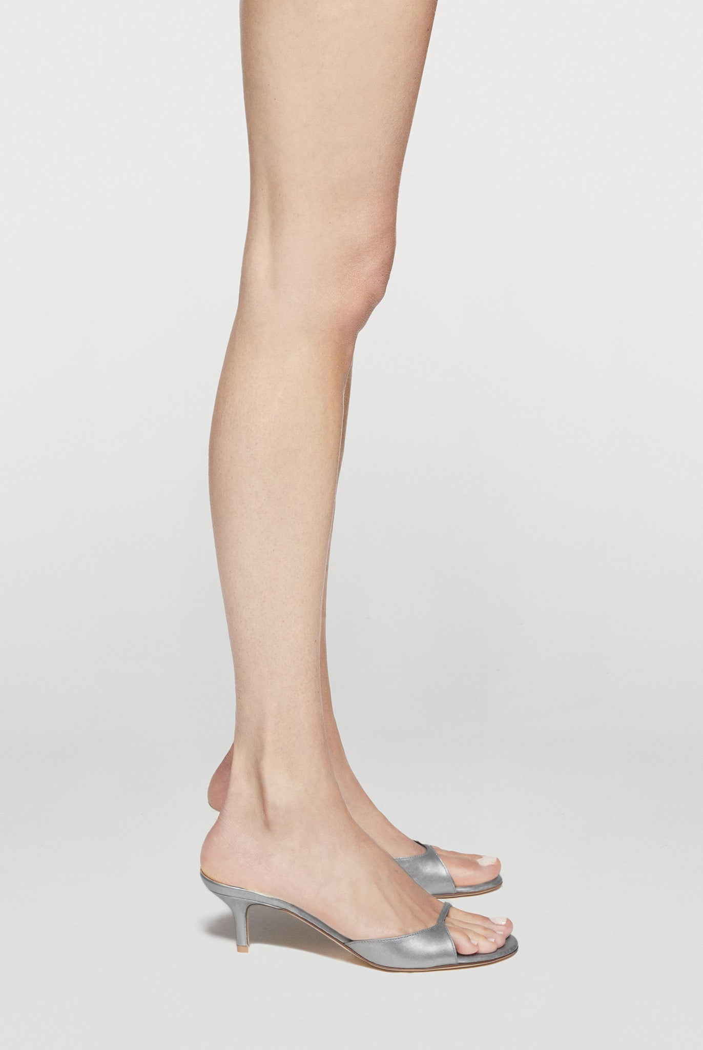 A pair of metallic silver mule sandals with kitten heels, shown on a model's feet against a light gray studio background.