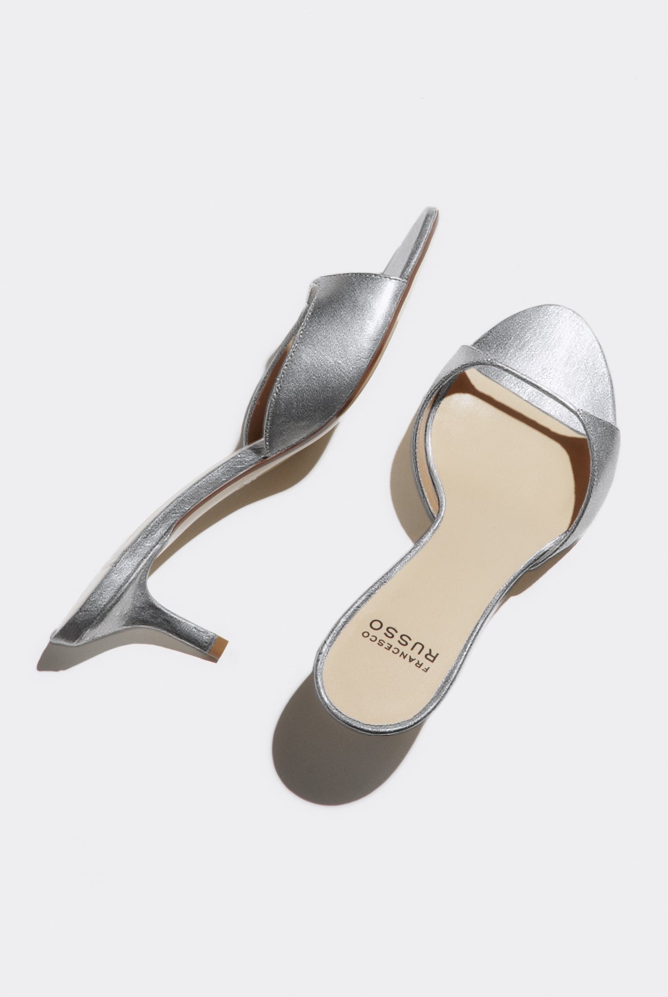 A pair of metallic silver slingback mule heels with kitten heels, shown from top and side angles on a light gray background. The brand name 'agency costume' is printed on the beige insole of the right shoe.