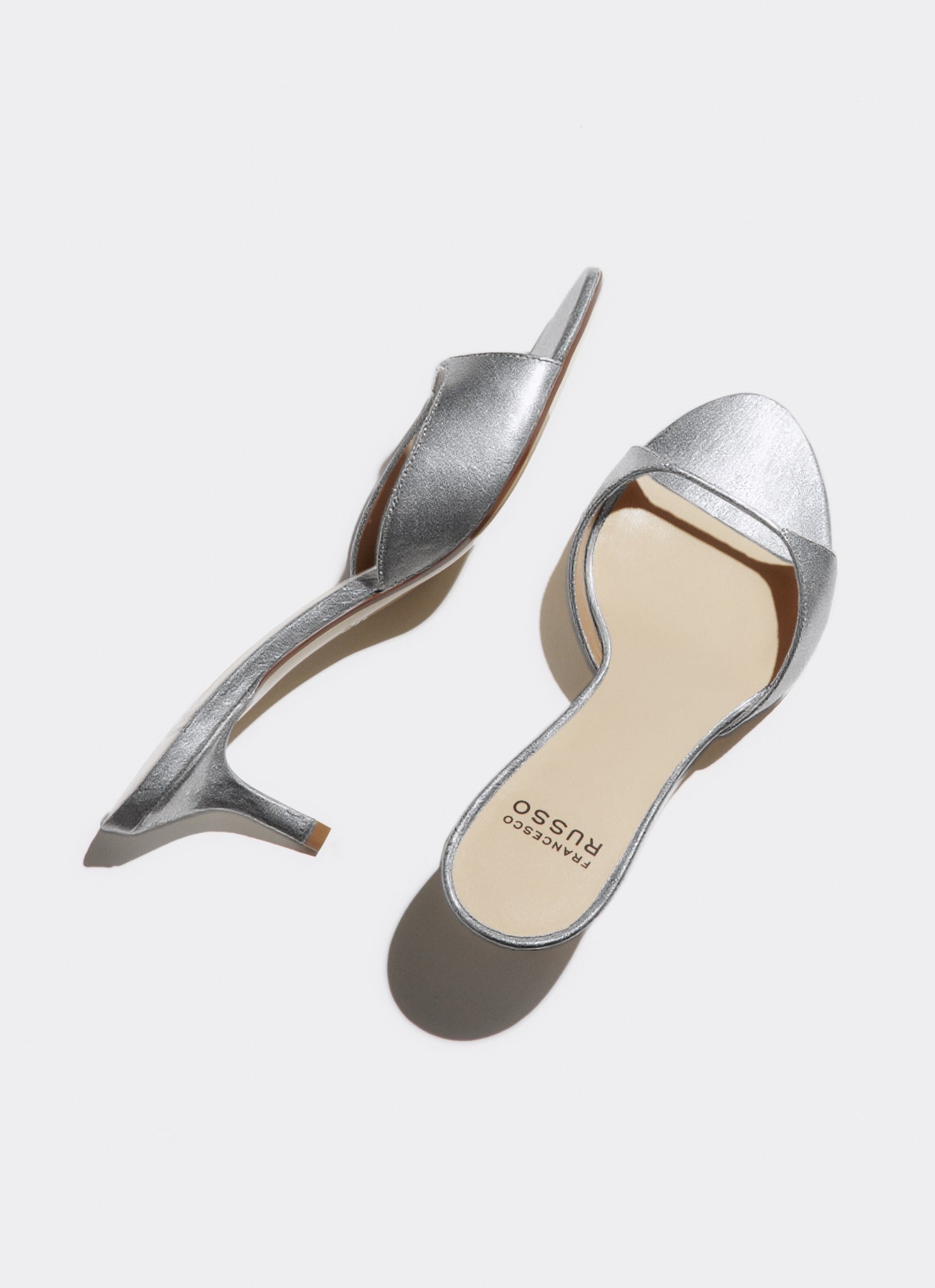A pair of metallic silver slingback mule heels with kitten heels, shown from top and side angles on a light gray background. The brand name 'agency costume' is printed on the beige insole of the right shoe.